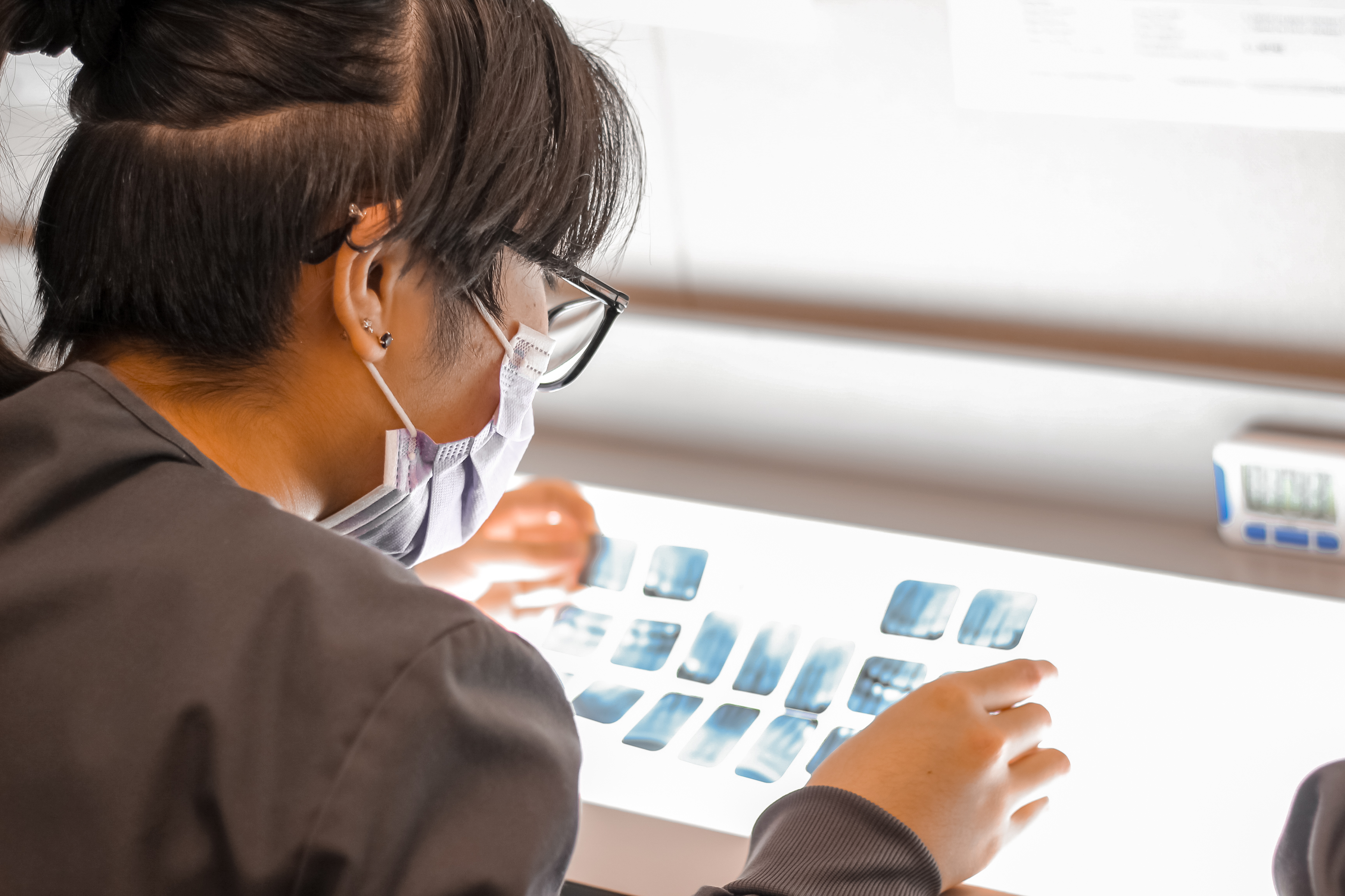 a person wearing a face mask and glasses, closely examining a series of X-ray images on a lightbox.